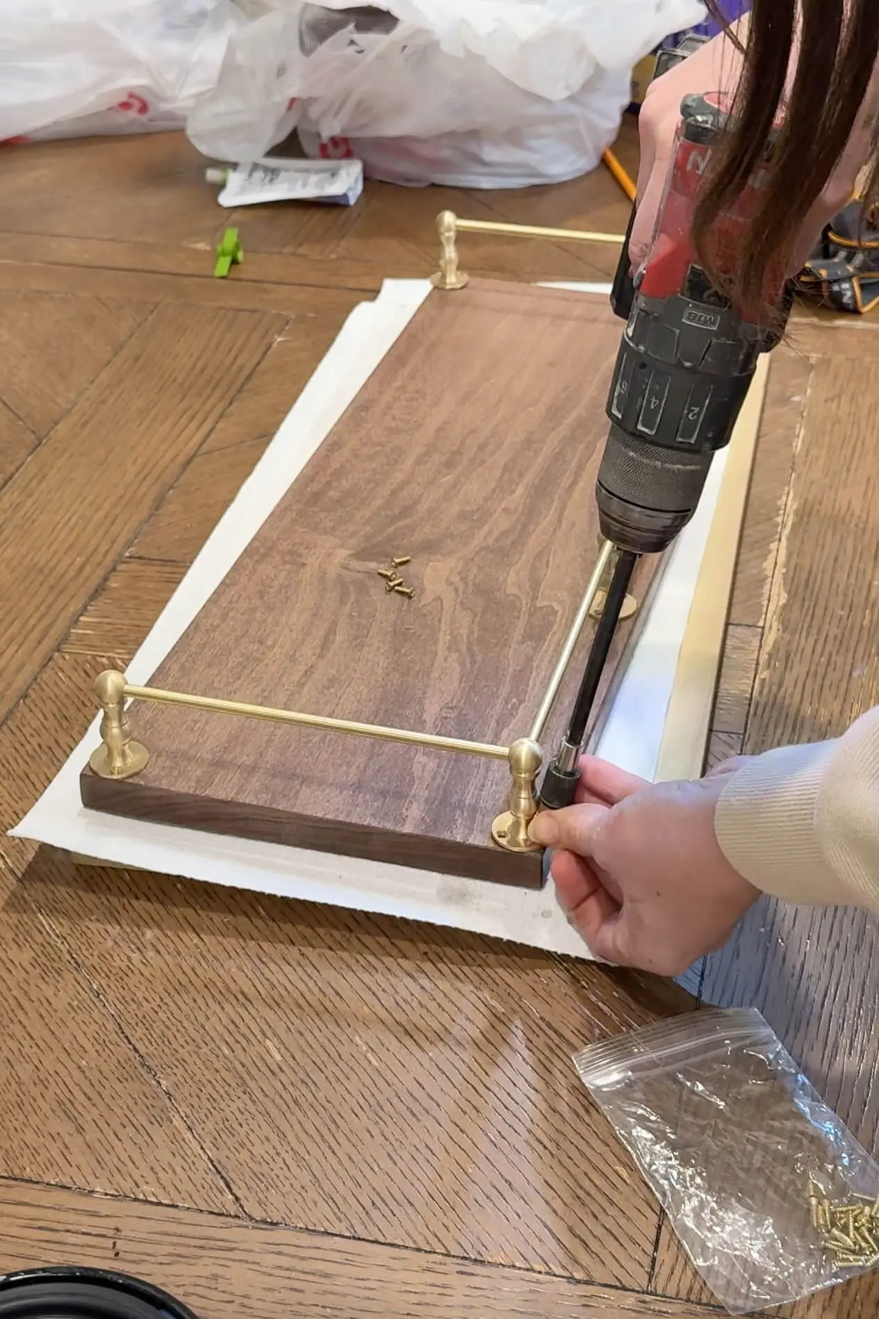 The final steps of assembling a wooden shelf as a woman secures a brass rod to the surface with a power drill, while screws and tools are scattered nearby.